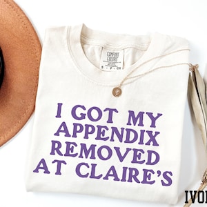 May include: A white t-shirt with the text "I GOT MY APPENDIX REMOVED AT CLAIRE'S" printed in purple. The shirt is laying flat on a white surface. The word "IVORY" is printed in the bottom right corner of the image.