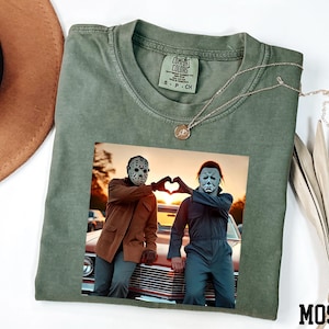 May include: Moss green Comfort Colors t-shirt featuring a graphic of two figures in Halloween masks making a heart shape with their hands. A silver necklace with a pendant is on the shirt.