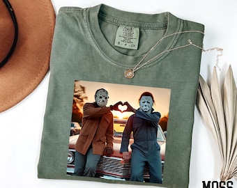 May include: Moss green Comfort Colors t-shirt featuring a graphic of two figures in Halloween masks making a heart shape with their hands. A silver necklace with a pendant is on the shirt.