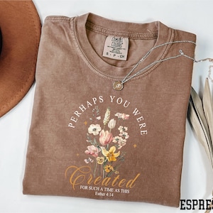 May include: Espresso-colored t-shirt with the text "Perhaps You Were Created" and a floral design. The shirt includes the Bible verse Esther 4:14. A silver necklace with a pendant is draped across the shirt.