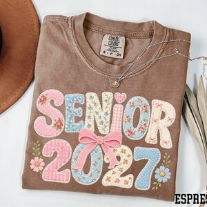 A brown t-shirt with the text "SENIOR 2027" printed on it, along with floral and heart-shaped decorations. The t-shirt is placed on a white surface, and there is a brown hat and some leaves nearby.