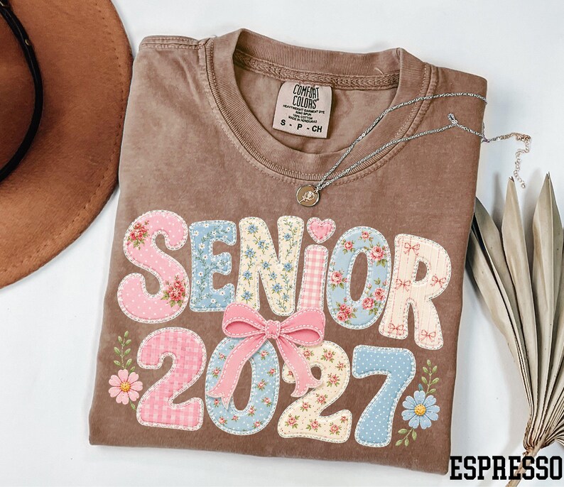 A brown t-shirt with the text "SENIOR 2027" printed on it, along with floral and heart-shaped decorations. The t-shirt is placed on a white surface, and there is a brown hat and some leaves nearby.