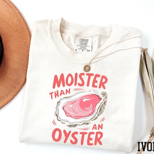 May include: A white t-shirt with the text "Moister than an oyster" in pink lettering. The shirt also features a cartoon illustration of an oyster with a pink interior. The shirt is labeled "Ivory" in the bottom right corner.