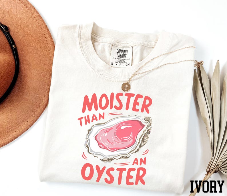May include: A white t-shirt with the text "Moister than an oyster" in pink lettering. The shirt also features a cartoon illustration of an oyster with a pink interior. The shirt is labeled "Ivory" in the bottom right corner.