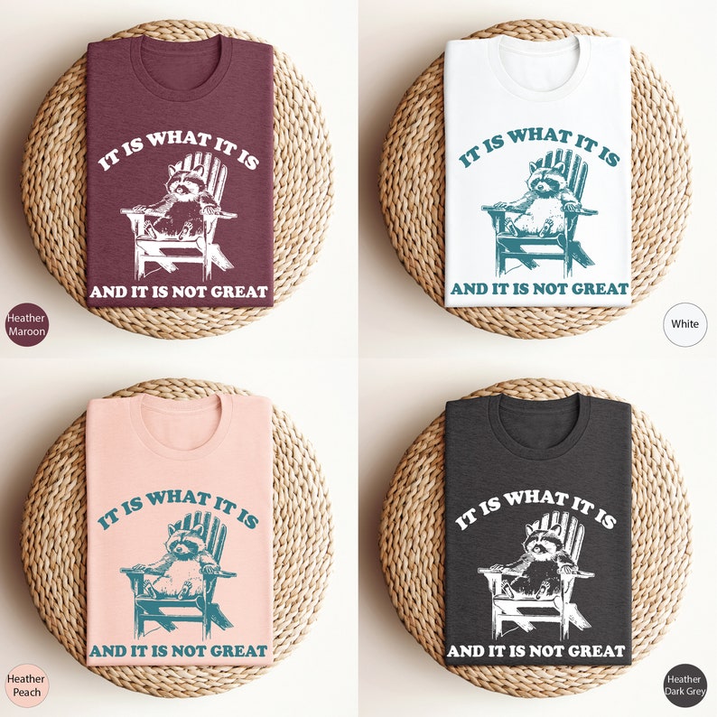 May include: Four t-shirts with a raccoon sitting in a chair and the text "It is what it is and it is not great". The t-shirts are in the colors heather maroon, white, heather peach, and heather dark grey.