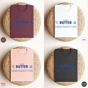 Puede incluir: Cuatro camisetas de diferentes colores, Heather Marr&oacute;n, Blanco, Heather Melocot&oacute;n y Heather Gris Oscuro, con un gr&aacute;fico de una barra de mantequilla con el texto "SALTED BUTTER AA" y una tabla que muestra el peso neto y la calidad de la mantequilla.