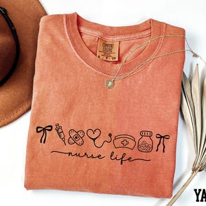 a pink t-shirt with a graphic design, a brown hat, a necklace, and a fan, all arranged on a white background.