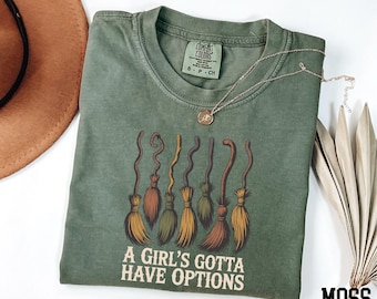 May include: A moss green t-shirt with a crew neck and the text "A Girl's Gotta Have Options" below a graphic of six stylized brooms. A gold necklace with a pendant is draped across the shirt. The word "MOSS" is in the bottom right corner.