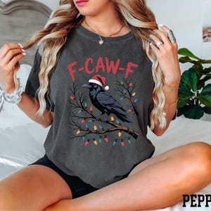 A woman wearing a gray t-shirt with a graphic design of a black crow wearing a Santa hat, sitting on a bed with a potted plant in the background.
