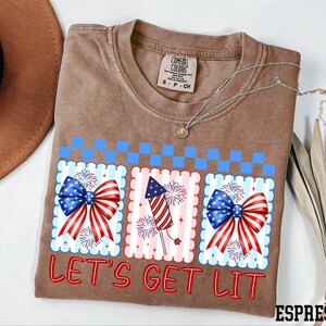 A t-shirt with a patriotic design, including a fireworks display and a ribbon, along with the text "Let's Get Lit" printed on it.