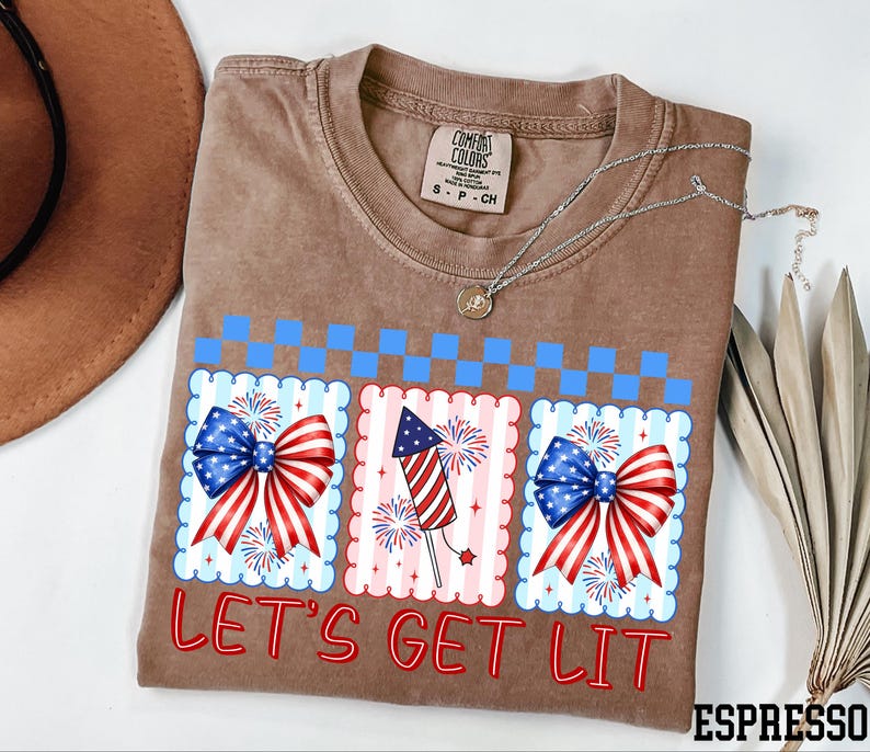 A t-shirt with a patriotic design, including a fireworks display and a ribbon, along with the text "Let's Get Lit" printed on it.