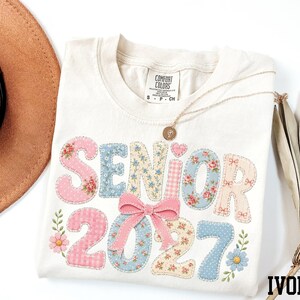a white t-shirt with the text "SENIOR 2027" printed on it, along with floral designs. The t-shirt is placed on a white surface, and there is a brown hat and a fan visible in the background.