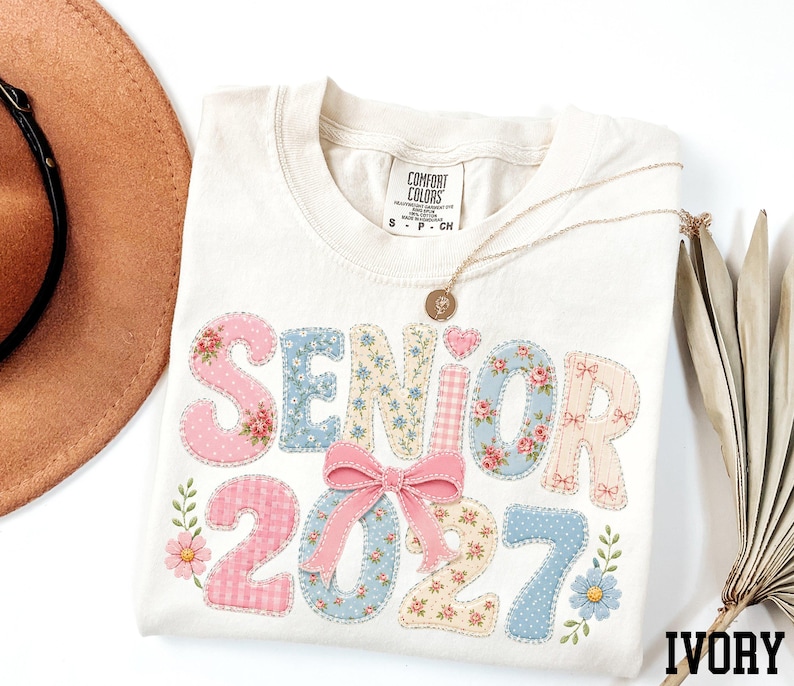 a white t-shirt with the text "SENIOR 2027" printed on it, along with floral designs. The t-shirt is placed on a white surface, and there is a brown hat and a fan visible in the background.