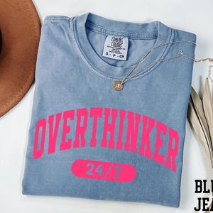 A blue t-shirt with the text "OVERTHINKER" printed in pink, along with the number "74/7" below it. The t-shirt is folded and placed on a white surface, accompanied by a brown hat and some leaves.