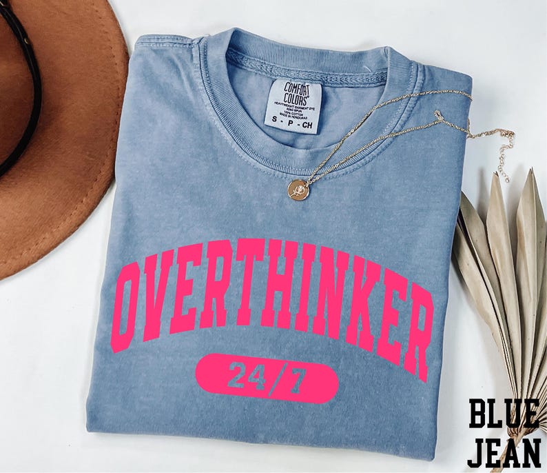 A blue t-shirt with the text "OVERTHINKER" printed in pink, along with the number "74/7" below it. The t-shirt is folded and placed on a white surface, accompanied by a brown hat and some leaves.