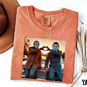 a t-shirt with a printed photo of two people posing with their hands in a heart shape, along with a necklace and a hat.
