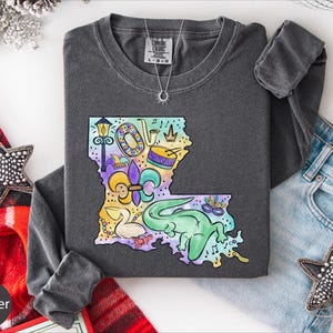 a gray long-sleeved shirt with a colorful, whimsical design of a map of Louisiana, surrounded by various decorative items such as a star, a scarf, and a necklace.