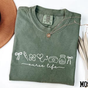 a green t-shirt with white text and illustrations, including a stethoscope, a heart, a pill bottle, and a ribbon, along with a necklace and a hat.