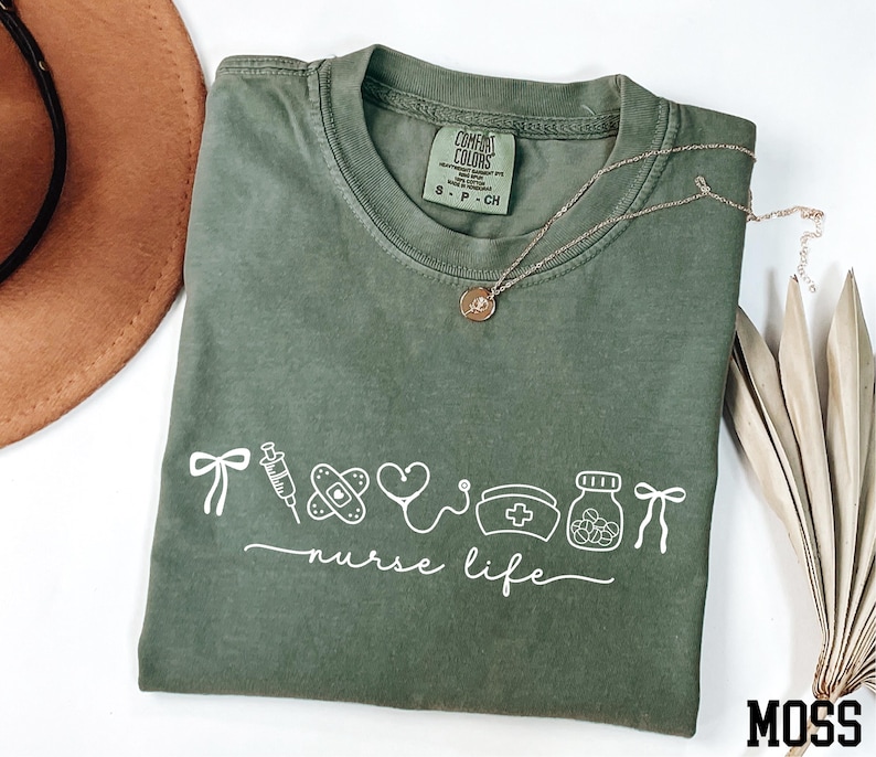 a green t-shirt with white text and illustrations, including a stethoscope, a heart, a pill bottle, and a ribbon, along with a necklace and a hat.