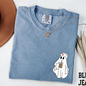 May include: A blue Comfort Colors t-shirt with a graphic of a ghost wearing a nurse's cap and holding an iced coffee. A gold necklace and the words "BLUE JEAN" are also visible.