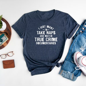 May include: A navy blue t-shirt with white text that reads "I just want to take naps and watch true crime documentaries". The shirt is laid out on a white surface with a pair of jeans, a pair of white sneakers, a wooden tray with a plant, a watch, and a camera.