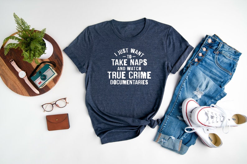 May include: A navy blue t-shirt with white text that reads "I just want to take naps and watch true crime documentaries". The shirt is laid out on a white surface with a pair of jeans, a pair of white sneakers, a wooden tray with a plant, a watch, and a camera.