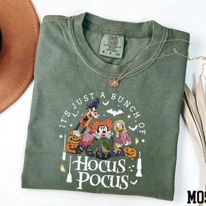 A green t-shirt with a Halloween-themed graphic design, including characters from the Disney movie "Hocus Pocus" and the text "It's just a bunch of Hocus Pocus".