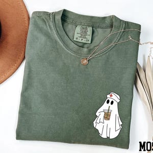a green t-shirt with a cartoon ghost wearing a nurse's hat, along with a brown hat, a necklace, and some dried leaves.