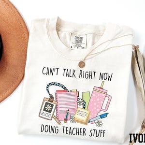 May include: Ivory-colored t-shirt with the text "Can't Talk Right Now Doing Teacher Stuff." The graphic includes a notepad, pencil, ruler, and a pink water bottle. A gold necklace is draped across the shirt.