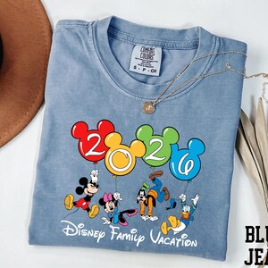 A blue t-shirt with a Disney-themed graphic and text, along with a hat and some decorative leaves, is displayed on a white surface.