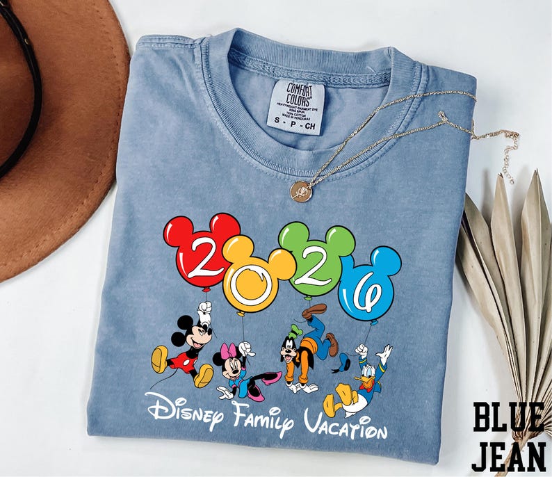 A blue t-shirt with a Disney-themed graphic and text, along with a hat and some decorative leaves, is displayed on a white surface.