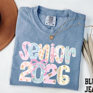 May include: A blue jean Comfort Colors t-shirt with the words "Senior 2026" in a floral and patterned design. A gold necklace with a pendant is draped across the shirt. The shirt is folded and displayed with a brown hat and dried plant.