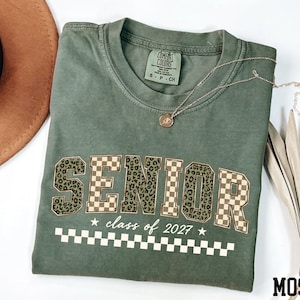 May include: An olive green crew neck t-shirt with the word SENIOR printed in a mix of leopard print and tan checkerboard patterns. Below the word, the text reads class of 2027 with two stars and a white checkerboard stripe at the bottom.