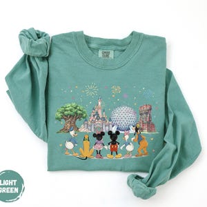 A green long-sleeved t-shirt with a printed design depicting Disney characters and landmarks, including Mickey Mouse, Donald Duck, Goofy, and Eiffel Tower.