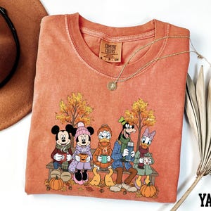 a red t-shirt with a cartoon illustration of Disney characters, including Minnie Mouse, Donald Duck, Goofy, and Mickey Mouse, sitting on a bench and holding cups of coffee. The t-shirt is displayed on a white background, along with a hat and a fan.
