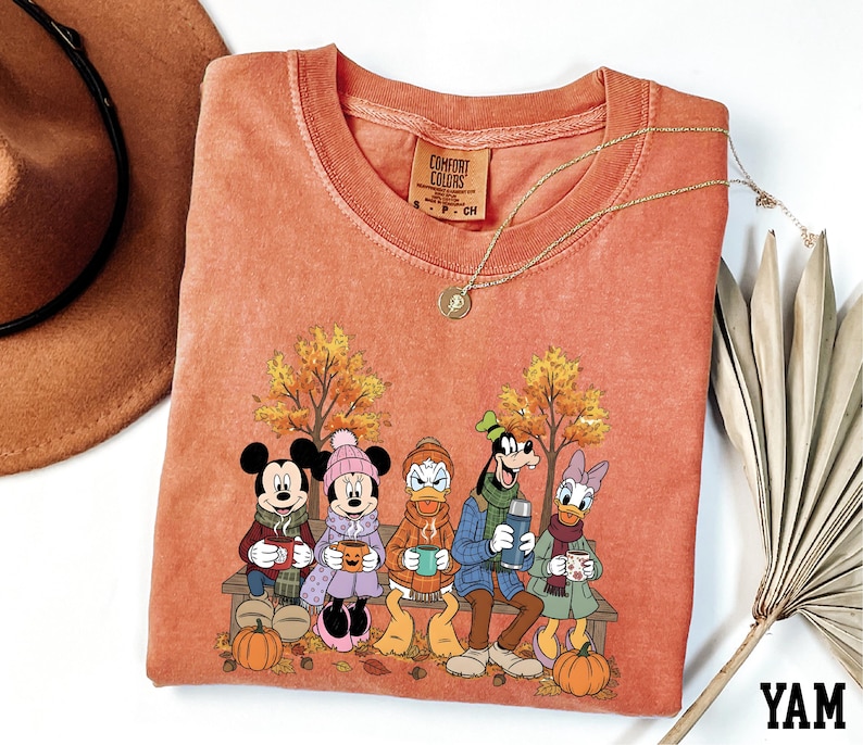 a red t-shirt with a cartoon illustration of Disney characters, including Minnie Mouse, Donald Duck, Goofy, and Mickey Mouse, sitting on a bench and holding cups of coffee. The t-shirt is displayed on a white background, along with a hat and a fan.