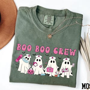 A green t-shirt with a graphic design of five ghost characters wearing nurse outfits and holding various Halloween-themed items, such as a pumpkin, spider web, and candy cane.