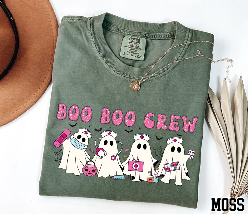 A green t-shirt with a graphic design of five ghost characters wearing nurse outfits and holding various Halloween-themed items, such as a pumpkin, spider web, and candy cane.