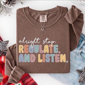 May include: Espresso-colored long-sleeve shirt with the text "alright stop. REGULATE AND LISTEN" in colorful block letters. A silver necklace with a sun pendant is on the shirt. The shirt is folded on a white surface with denim jeans and holiday decorations.