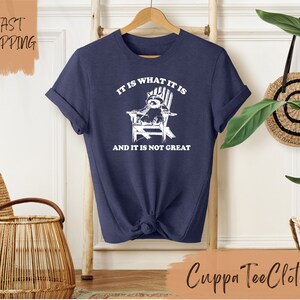 May include: A navy blue t-shirt with a white graphic of a raccoon sitting in a chair. The text "It is what it is and it is not great" is printed on the shirt.