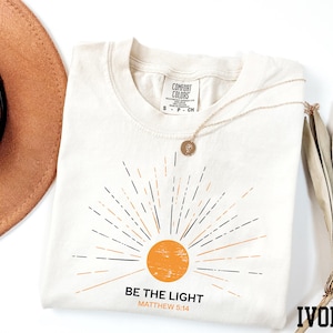 May include: Ivory-colored t-shirt with a sunburst design in orange and black. The text "BE THE LIGHT" and "MATTHEW 5:14" are printed below the sun graphic. A gold necklace with a pendant is draped on the shirt.