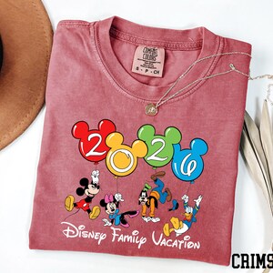 A red t-shirt with a Disney-themed graphic and text, along with a necklace and a hat, is displayed on a white surface.