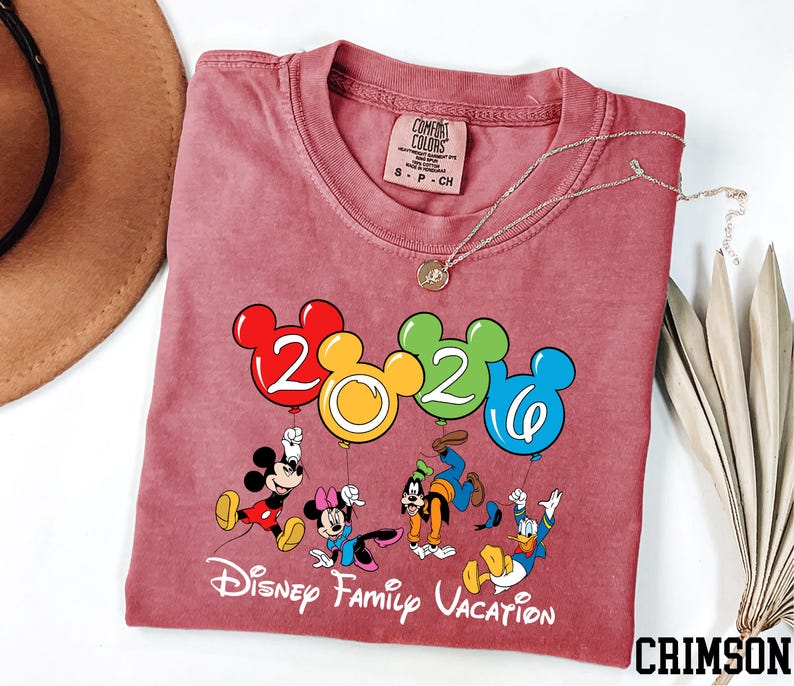 A red t-shirt with a Disney-themed graphic and text, along with a necklace and a hat, is displayed on a white surface.