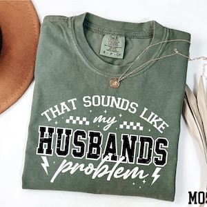 May include: A moss green t-shirt with the text "THAT SOUNDS LIKE my HUSBANDS problem" in white and black. The shirt has a crew neck and short sleeves. A gold necklace is draped across the shirt.