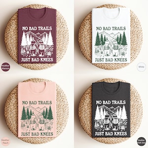 Może przedstawiać: Cztery koszulki z nadrukiem graficznym przedstawiającym dwa niedźwiedzie wędrujące po lesie z tekstem "No Bad Trails Just Bad Knees". Koszulki są dostępne w czterech kolorach: melanż brązowy, biały, melanż brzoskwiniowy i melanż ciemnoszary.