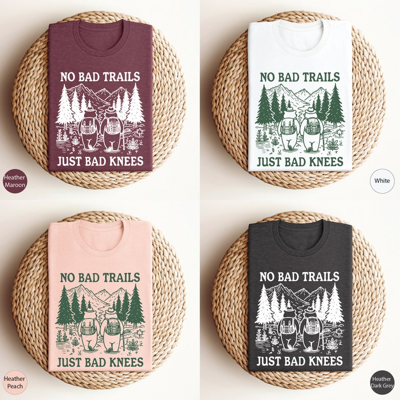 Może przedstawiać: Cztery koszulki z nadrukiem graficznym przedstawiającym dwa niedźwiedzie wędrujące po lesie z tekstem "No Bad Trails Just Bad Knees". Koszulki są dostępne w czterech kolorach: melanż brązowy, biały, melanż brzoskwiniowy i melanż ciemnoszary.
