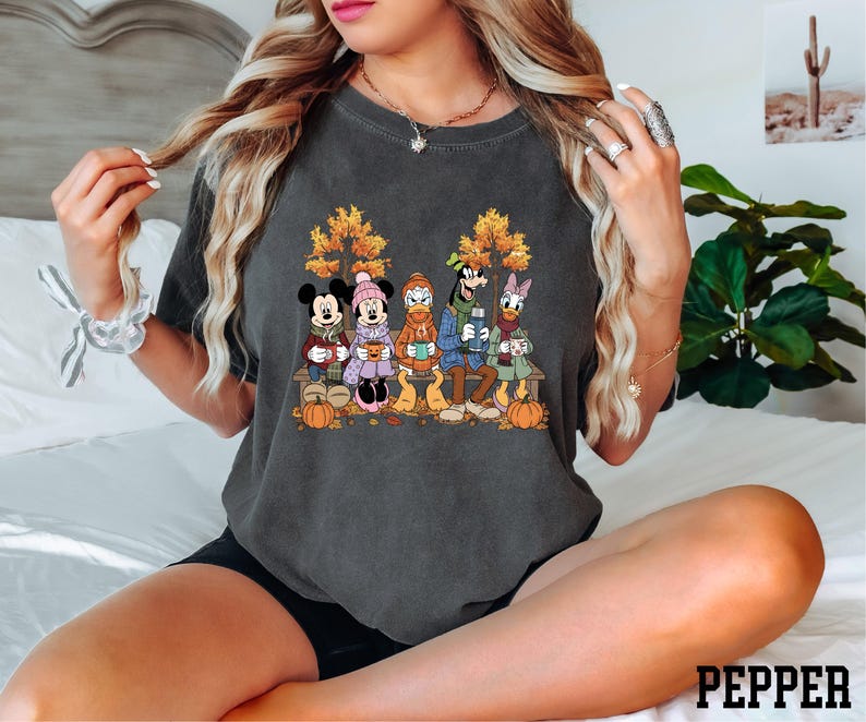 A woman wearing a t-shirt with a graphic of Disney characters is sitting on a bed.