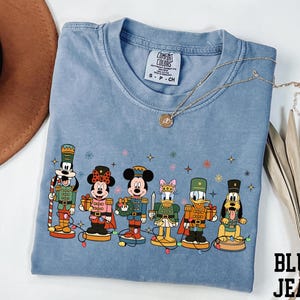 May include: A blue Comfort Colors t-shirt with a graphic of Disney characters as nutcrackers. Mickey, Minnie, Donald, and Goofy are depicted holding gifts. The shirt has a crew neck and short sleeves. The text "BLUE JEAN" is visible in the bottom right corner.
