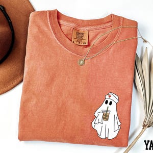 a red t-shirt with a cartoon ghost wearing a nurse's hat and holding a drink, along with a brown hat and a fan.