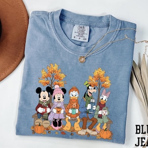 A blue t-shirt with a cartoon image of Disney characters Mickey Mouse, Minnie Mouse, Donald Duck, and Goofy sitting on a bench with autumn leaves and pumpkins.